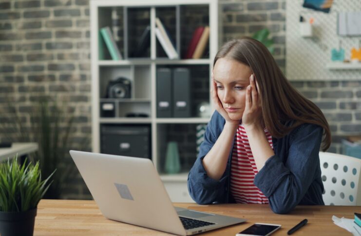 A woman sitting with a laptop in front of her, with her head in her hands, looking overworked.