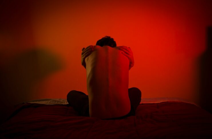 A shirtless man sits on a bed with his back to the camera.