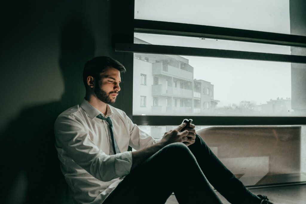 A sad man sitting on the floor of an office while looking at his phone.