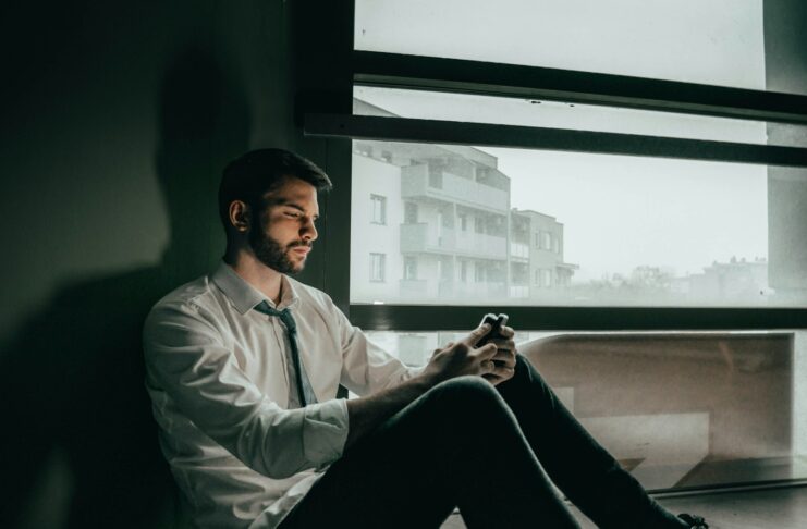 A sad man sitting on the floor of an office while looking at his phone.