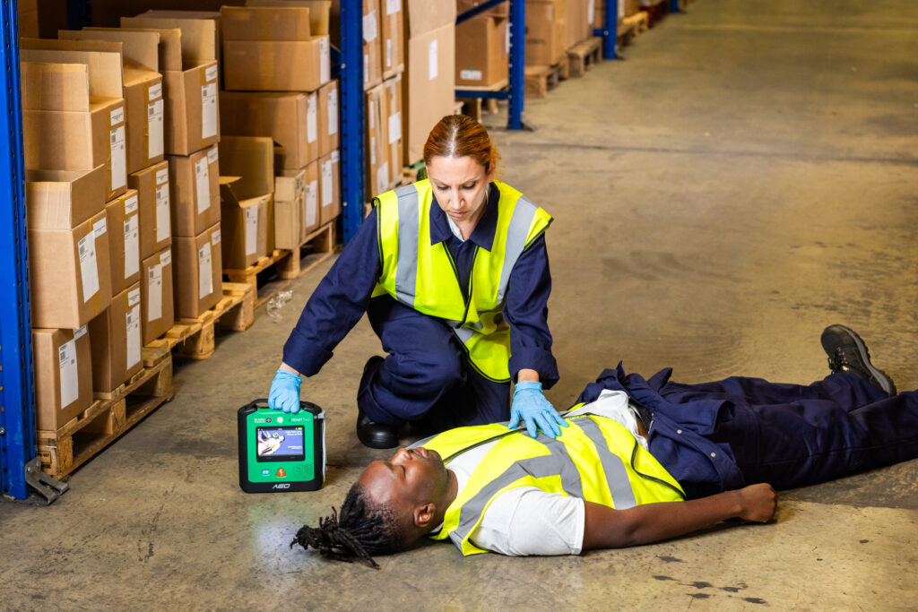 A woman uses a defibrilator on a man lying on the ground in a warehouse.