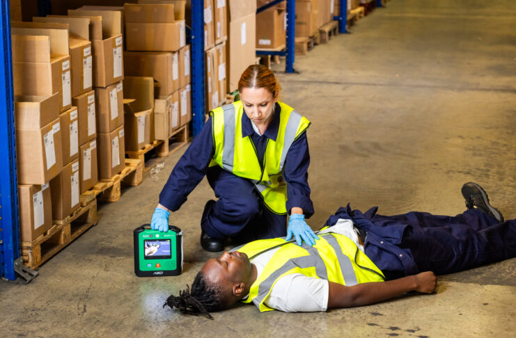 A woman uses a defibrilator on a man lying on the ground in a warehouse.