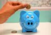 Midlife Money Squeeze ‘Leaves Employees Managing Financial Pressure in Silence’ Hand inserting a coin into a blue piggy bank for savings and money management.