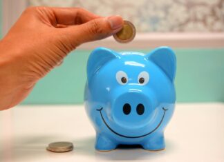 Midlife Money Squeeze ‘Leaves Employees Managing Financial Pressure in Silence’ Hand inserting a coin into a blue piggy bank for savings and money management.
