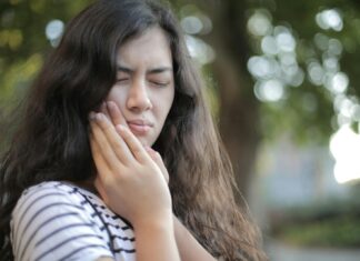 Workers Hide Dental Pain as Stigma and ‘Push Through’ Culture Take Hold Portrait of a young woman feeling toothache, showing discomfort outdoors with a pained facial expression.