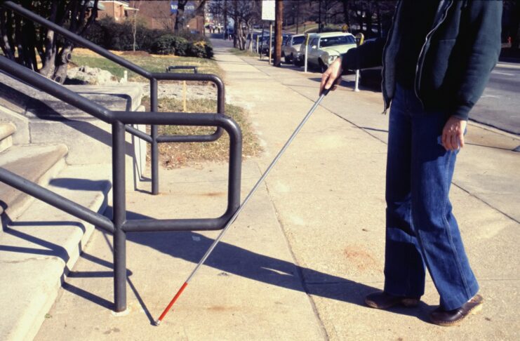 A blind man walks on a street with the aid of a stick.