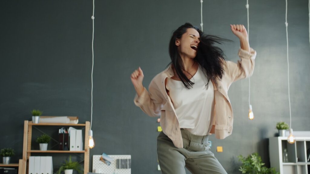 A woman joyfully dancing in an office with arms raised.