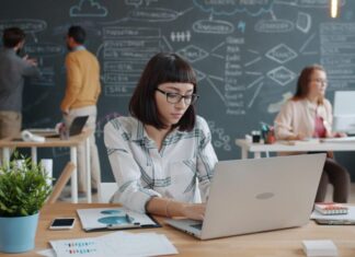 Young Employees ‘Call for Greater Workplace Wellbeing Support’ Woman working on laptop in modern office with team.
